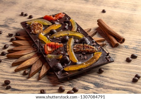 Delicious black chocolate with candied fruits on a wooden stand on a wooden background with a cinnamon stick and grains of coffee.