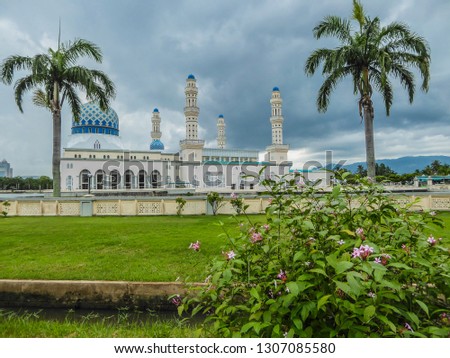 Masjid Bandaraya in Kota Kinabalu, Sabah, Malaysia. Lush green lawn in front of the mosque. Two palm trees on the sides of the temple. A flower bush in front. Huge overcast, storm is coming.