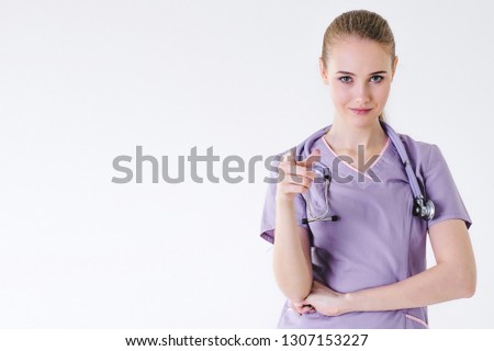Woman doctor blonde young light gray background studio day beautiful one looks with a smile surprised rejoices thumbs up. Indicates when meeting approves the selection.