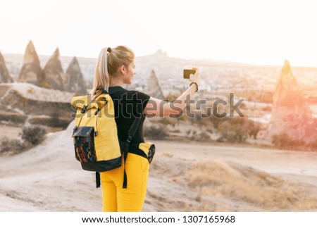 Blond hair woman with yellow authentic backpack taking landscape nature photo with phone. Travel adventures in desert. Goreme, Cappadocia, Turkey.  Copy space background, empty place for text.