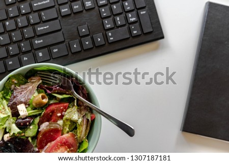 Healthy veggie bowl with green salad, tomatoes, green and black olives, feta cheese. Keyboard and notepad in the background.