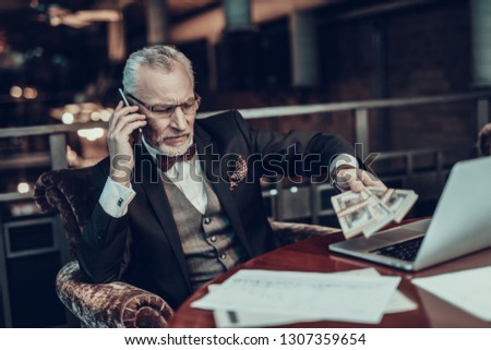 Businessman Using Laptop. Old Businessman . Man is Wearing in Black Suit. Experienced Entrepreneur. Strict Man. looking at Laptop. Working Late. talking on Phone. holding stack of money