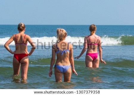 Three beautiful young women standing back in bikini in waves of ocean