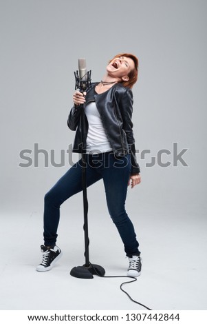 Full-length portrait of rock singer wearing leather jacket and keeping static mic, sings a song loudly on grey background. Concept of rock music and rave
