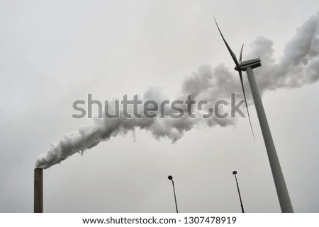 Wind turbine against a smoking industrial chimney of a power plant polluting the air of a grey sky at dawn.