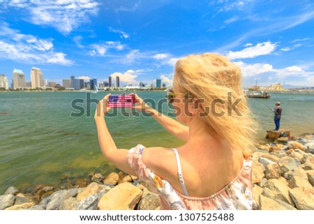 Blonde tourist takes photo of San Diego By skyline by mobile phone with American flag cover. Female lifestyle takes pictures in California summer holidays with her smartphone from Coronado Island, USA