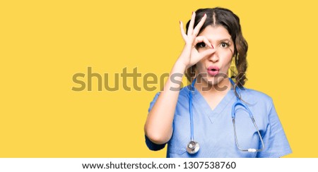 Young adult doctor woman wearing medical uniform doing ok gesture shocked with surprised face, eye looking through fingers. Unbelieving expression.