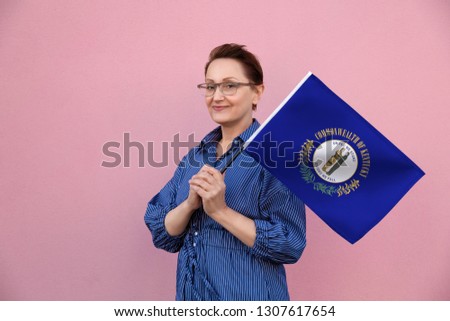 Kentucky flag. Woman holding Kentucky state flag. Nice portrait of middle aged lady 40 50 years old holding a large state flag over pink wall background on the street outdoor.