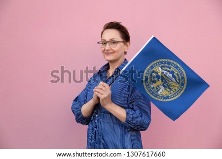 Nebraska flag. Woman holding Nebraska state flag. Nice portrait of middle aged lady 40 50 years old holding a large state flag over pink wall background on the street outdoor.