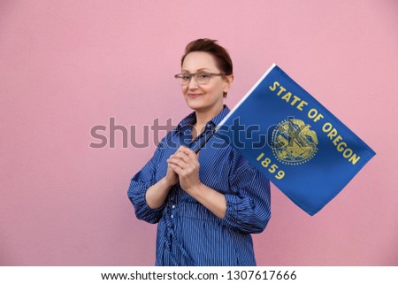 Oregon flag. Woman holding Oregon state flag. Nice portrait of middle aged lady 40 50 years old holding a large state flag over pink wall background on the street outdoor.