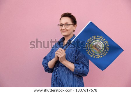 New Hampshire flag. Woman holding New Hampshire state flag. Nice portrait of middle aged lady 40 50 years old holding a large state flag over pink wall background on the street outdoor.