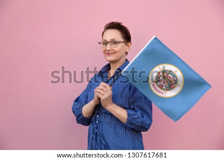 Minnesota flag. Woman holding Minnesota state flag. Nice portrait of middle aged lady 40 50 years old holding a large state flag over pink wall background on the street outdoor.
