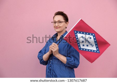 Arkansas flag. Woman holding Arkansas state flag. Nice portrait of middle aged lady 40 50 years old holding a large state flag over pink wall background on the street outdoor.
