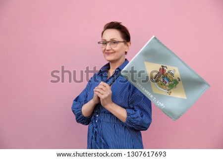 Delaware flag. Woman holding Delaware state flag. Nice portrait of middle aged lady 40 50 years old holding a large state flag over pink wall background on the street outdoor.