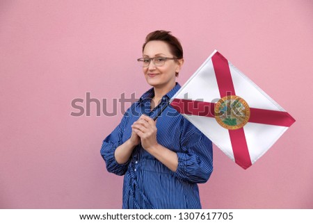 Florida flag. Woman holding Florida state flag. Nice portrait of middle aged lady 40 50 years old holding a large state flag over pink wall background on the street outdoor.