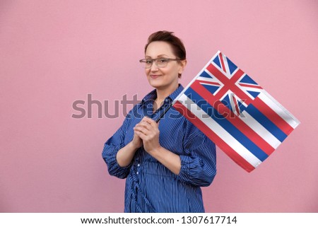 Hawaii flag. Woman holding Hawaiian state flag. Nice portrait of middle aged lady 40 50 years old holding a large state flag over pink wall background on the street outdoor.