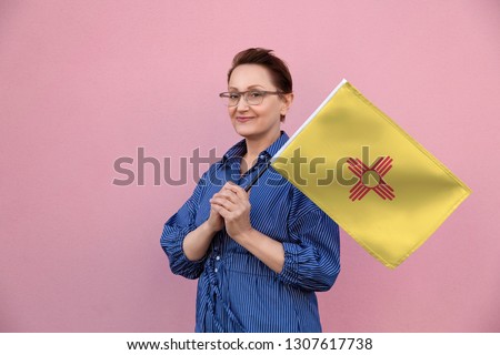 New Mexico flag. Woman holding New Mexico state flag. Nice portrait of middle aged lady 40 50 years old holding a large state flag over pink wall background on the street outdoor.