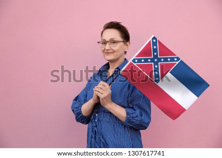 Mississippi flag. Woman holding Mississippi state flag. Nice portrait of middle aged lady 40 50 years old holding a large state flag over pink wall background on the street outdoor.