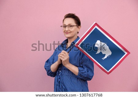 Wyoming flag. Woman holding Wyoming state flag. Nice portrait of middle aged lady 40 50 years old holding a large state flag over pink wall background on the street outdoor.
