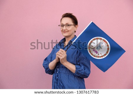 Virginia flag. Woman holding Virginia state flag. Nice portrait of middle aged lady 40 50 years old holding a large state flag over pink wall background on the street outdoor.