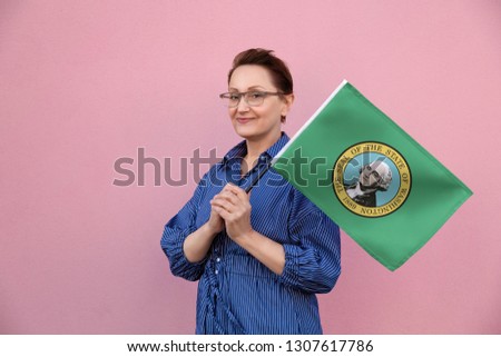 Washington flag. Woman holding Washington state flag. Nice portrait of middle aged lady 40 50 years old holding a large state flag over pink wall background on the street outdoor.