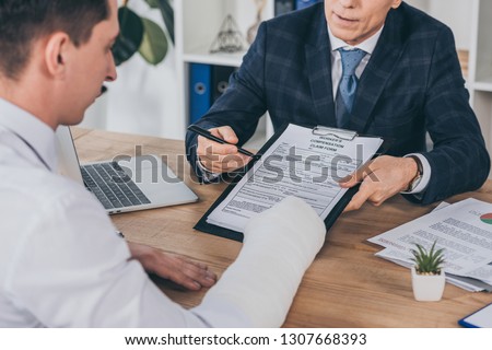 businessman in blue jacket giving form for compensation claim to worker with broken arm at table in office, compensation concept Royalty-Free Stock Photo #1307668393
