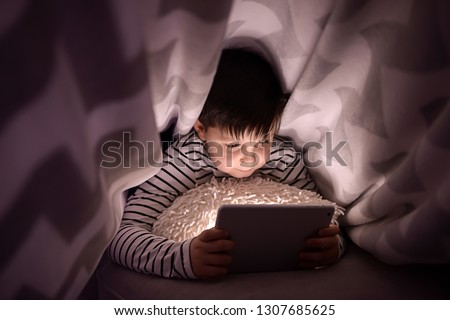 Cute little boy with tablet computer watching cartoons under blanket