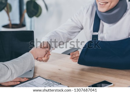cropped view of woman shaking hands with worker in neck brace and arm bandage over table in office, compensation concept