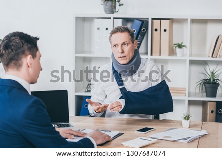 middle aged worker in neck brace with broken arm sitting at table and talking to businessman in blue jacket in office, compensation concept Royalty-Free Stock Photo #1307687074