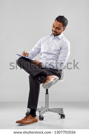 business, office worker and people concept - smiling indian businessman writing to clipboard over grey background