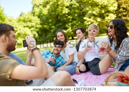 friendship, leisure and technology concept - guy taking picture on smartphone of friends drinking non alcoholic drinks at picnic in summer park