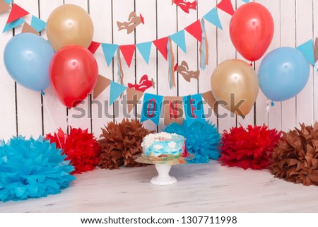 Photo zone with paper garlands, balloons, paper balls, pom poms, confetti and cream cake. Birthday cake. Smash cake. One year. Blue, gold, beige, brown colors. Pilot, Sky