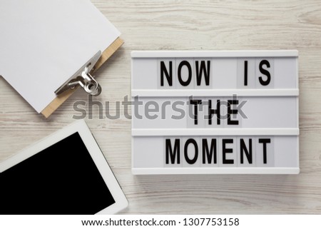 Modern board with text 'Now is the moment', tablet with blank screen, noticepad on a white wooden background, overhead view. From above, flat lay, top view. 
