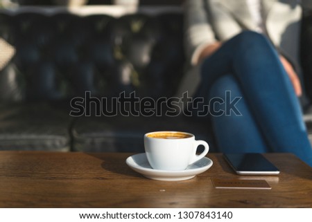 Close up photo of white cup of coffee, plastic card and portable telephone equipment on wooden table against blurred lady, sitting inside loft interior space in restaurant