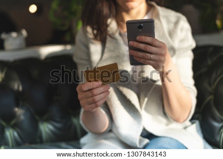Close up cropped photo of unrecognizable lady in trend, trendy clothes inside loft interior space in restaurant. She sitting in chair holding plastic card and portable telephone equipment