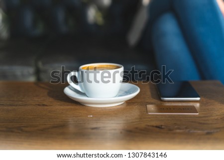 Selective focus and close up photo of white mug with drink, portable telephone equipment and credit card against blurred woman, sitting inside loft interior space in restaurant