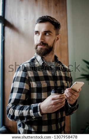 Bearded attractive man, handsome hipster bearded guy holding mobile cell phone and cup of morning coffee in new modern coffee shop via the searching network. Free wi-fi and technologies concept