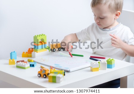 Little blond child draws at the table. Little boy sits on a chair at the white table. Boy playing with lots of colorful plastic blocks and toy train.