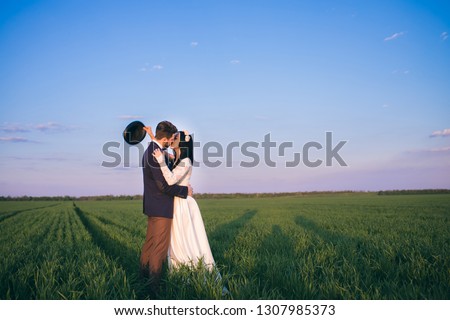 Fashionable bride and beautiful bride hugging in the field, sunset and hat in hand, the guy kisses the girl