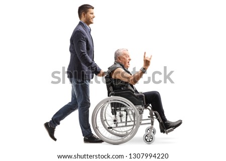Full length profile shot of a young man pushing an man in leather vest making a rock and roll hand sign in a wheelchair isolated on white background