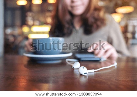 Closeup image of a woman drinking coffee while using mobile phone to listen music with earphone on wooden table in cafe
