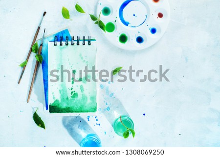 Artist workplace with tools and plants, green and blue watercolor sketchbooks, brushes and color swatches on a white background. Spring header with copy space