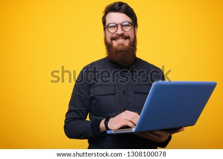 Picture of handsome young bearded man over yellow wall background isolated. Looking at camera using laptop. 