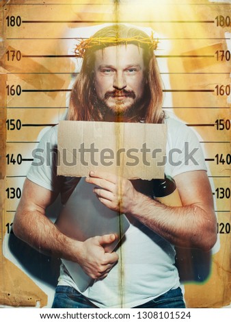 Male criminal suspect in the image of Jesus holds a laptop and a bottle of alcohol in his hand, is photographed against the background of the growth wall. Creative religious concept