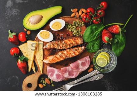 Keto, ketogenic diet, low carbohydrate content. Grilled salmon, vegetables, strawberries, cheese, ham and water with lemon. Black stone background. Top view Royalty-Free Stock Photo #1308103285