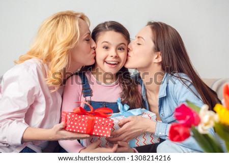 Two women and girl together at home celebration sitting on sofa granny and mom kissing child holding present box smiling surprised