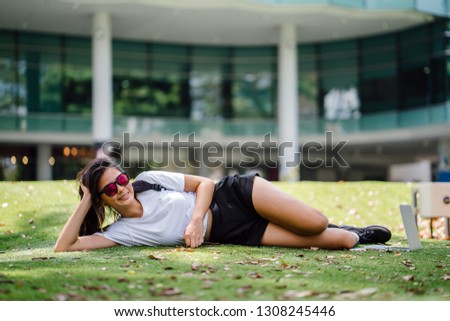 A beautiful and young Chinese Asian student is relaxing on the grass lawn on her university campus in the sun. She is lying on the grass on her side and is smiling. She is dressed casually. 