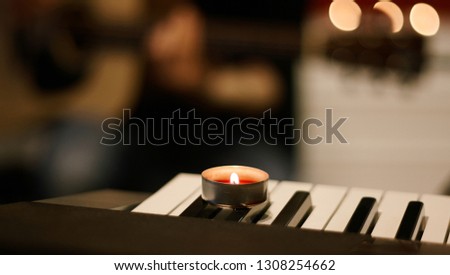 A burning candle with red wax stands on a musical synthesizer, and in the background a man plays the guitar