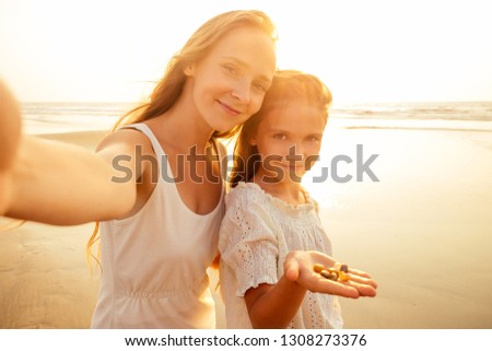 Happy stylishly mother and daughter taking selfie at sandy beach shell on a sunset.mothers Day.little girl blonde and beautiful woman taking pictures on the phone tourism abroad online video calling