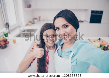 Self-portrait of two nice cute lovely sweet attractive charming cheerful mama wearing apron pre-teen girl showing thumbup advert ad in light white interior indoors